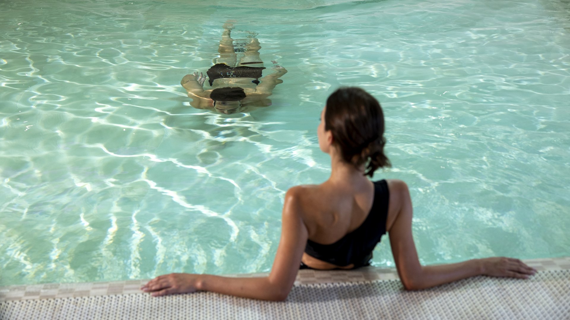A day dedicated to wellness Woman in black swimsuit relaxing at pool edge with another person swimming
